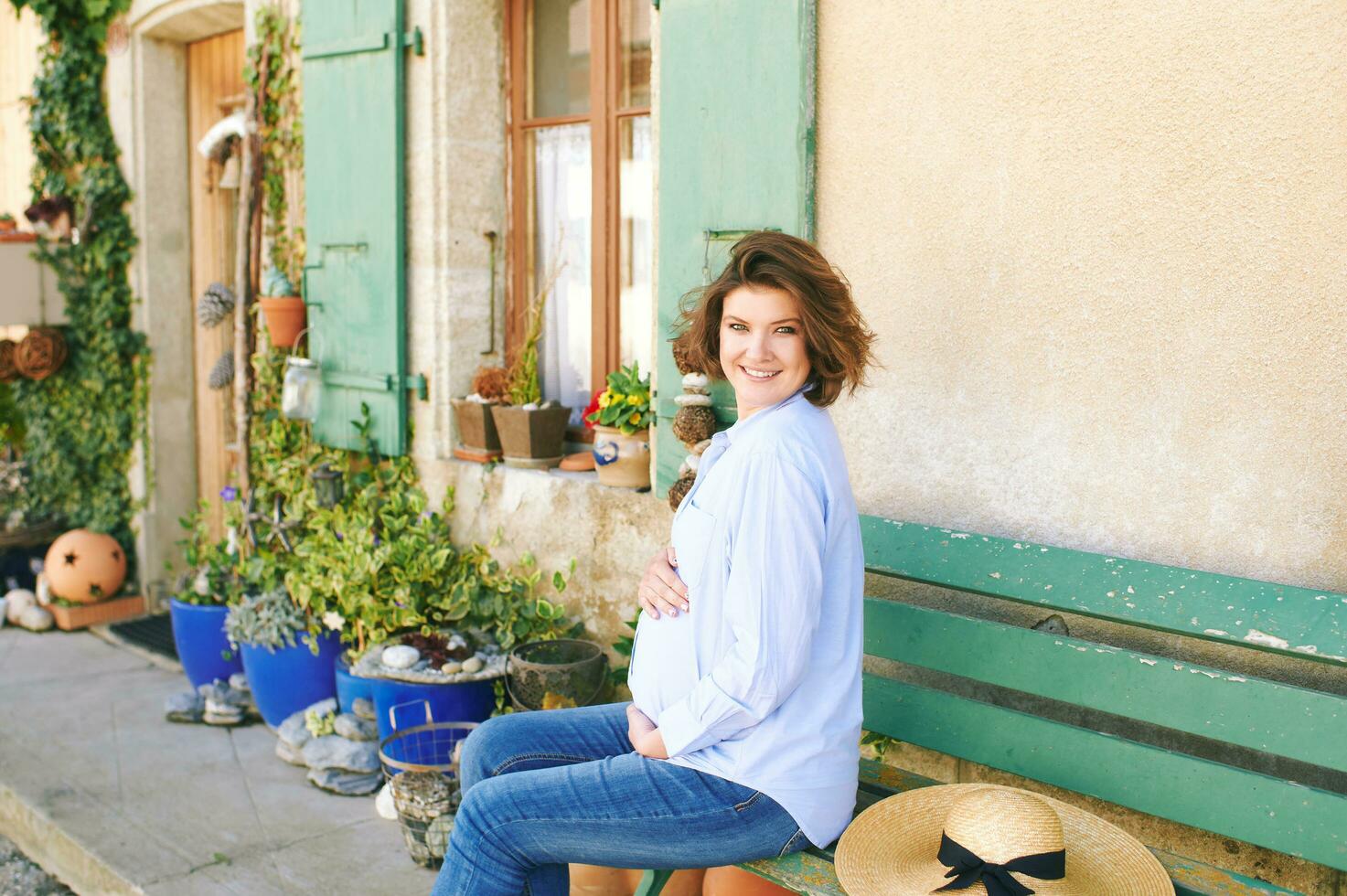 ung gravid kvinna Sammanträde på bänk Nästa till skön by hus Fasad foto