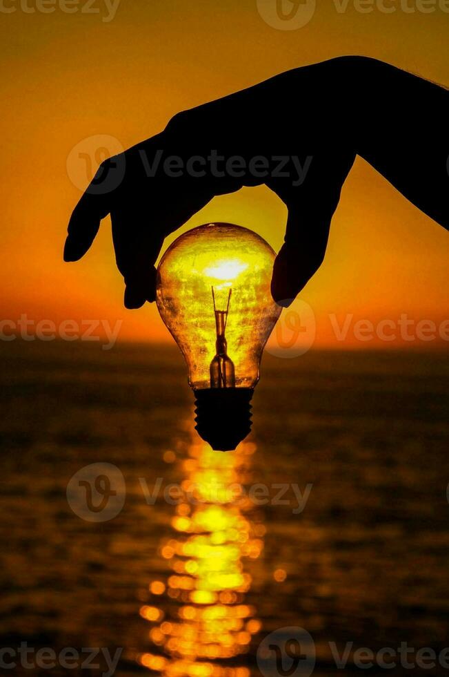 en person innehav en ljus Glödlampa över de hav på solnedgång foto