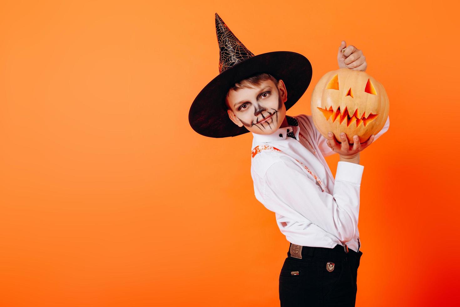 porträtt av pojke i djävulen maskerad smink och hatt som visar en pumpa. halloween koncept foto