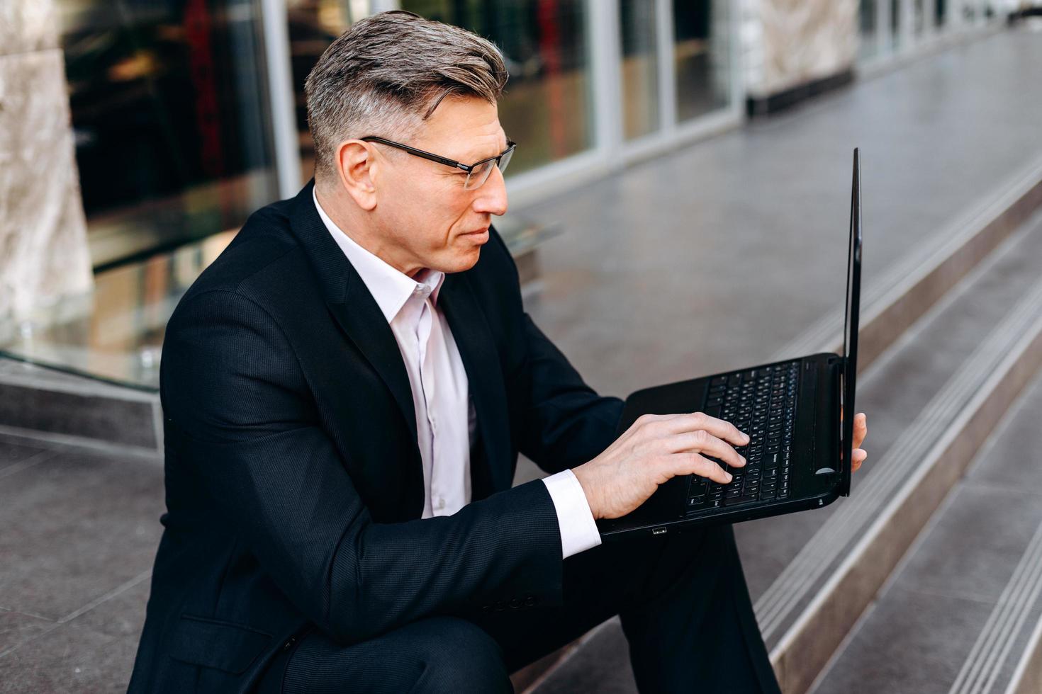 senior affärsman sitter på trottoaren, håller en bärbar dator och skriver. - bild foto
