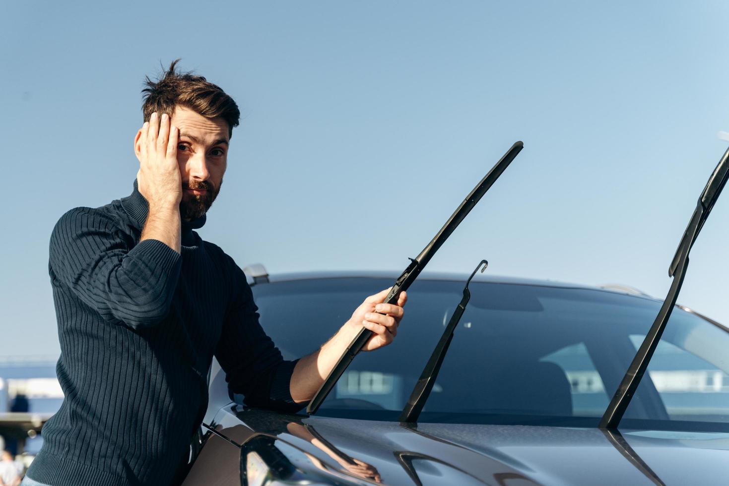 Herregud. kaukasisk man håller i huvudet med chockade känslor medan han byter vindrutetorkare på en bil. transport koncept foto