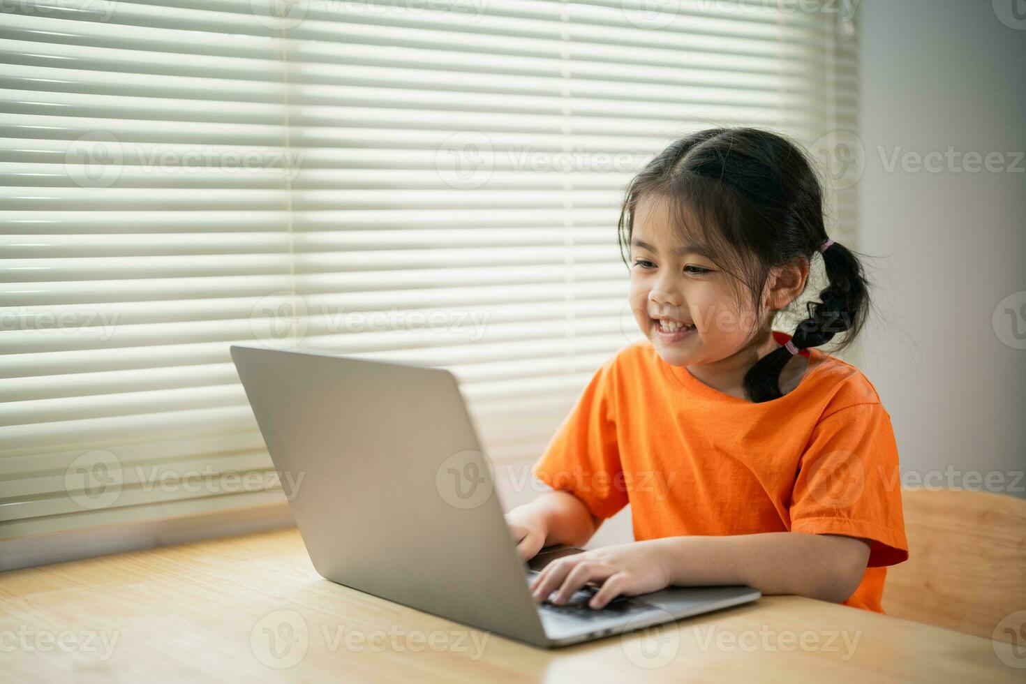 asiatisk bebis flicka bär t-shirt leende koncentrera till använda sig av bärbar dator och studie uppkopplad på trä tabell skrivbord i levande rum på Hem på natt. utbildning inlärning uppkopplad från Hem begrepp. foto