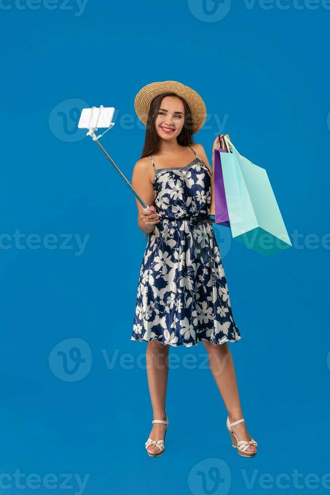 ung kvinna i tillfällig kläder med handla påsar använder sig av selfie pinne till ta en själv porträtt på blå studio bakgrund med kopia Plats foto