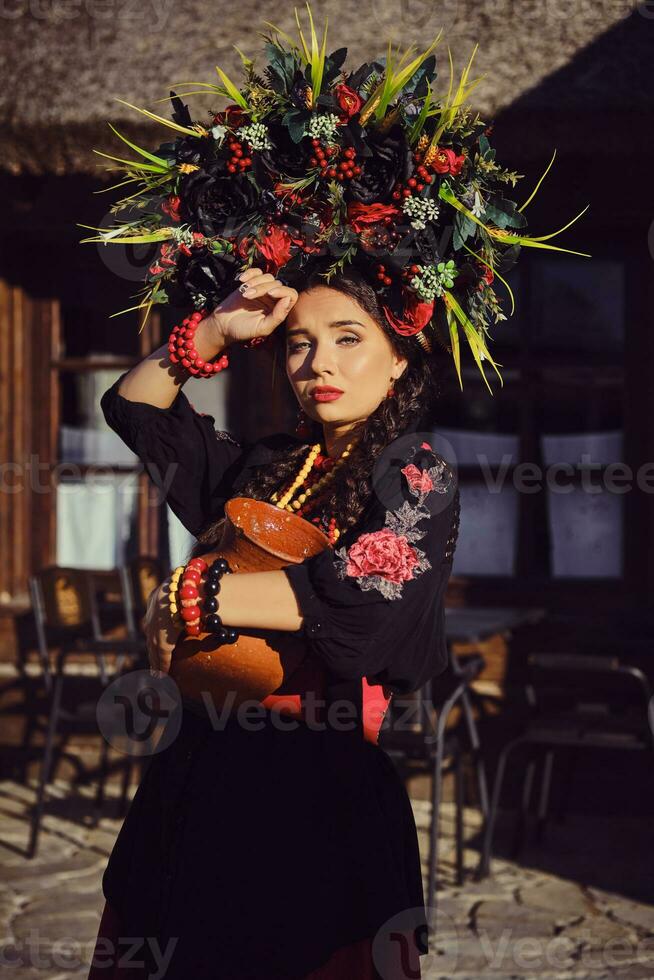 brunett flicka i en svart och röd ukrainska broderad äkta nationell kostym och en krans av blommor är Framställ stående på de Port. foto