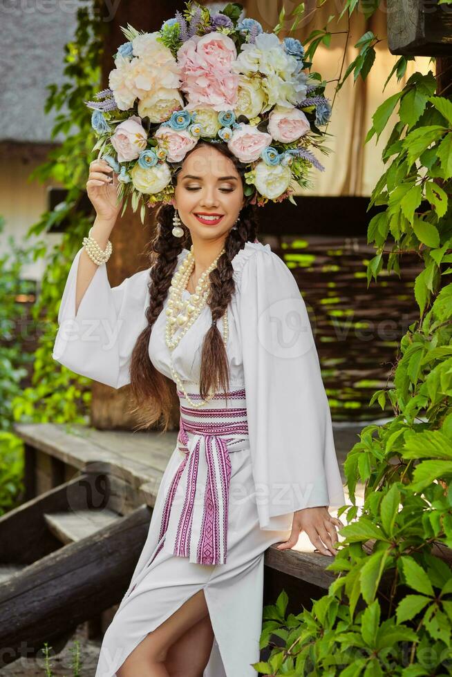 brunett flicka i en vit ukrainska äkta nationell kostym och en krans av blommor är Framställ mot en grön gård. närbild. foto