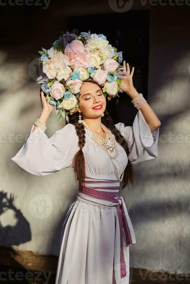 brunett flicka i en vit ukrainska äkta nationell kostym och en krans av blommor är Framställ mot en vit hydda. närbild. foto