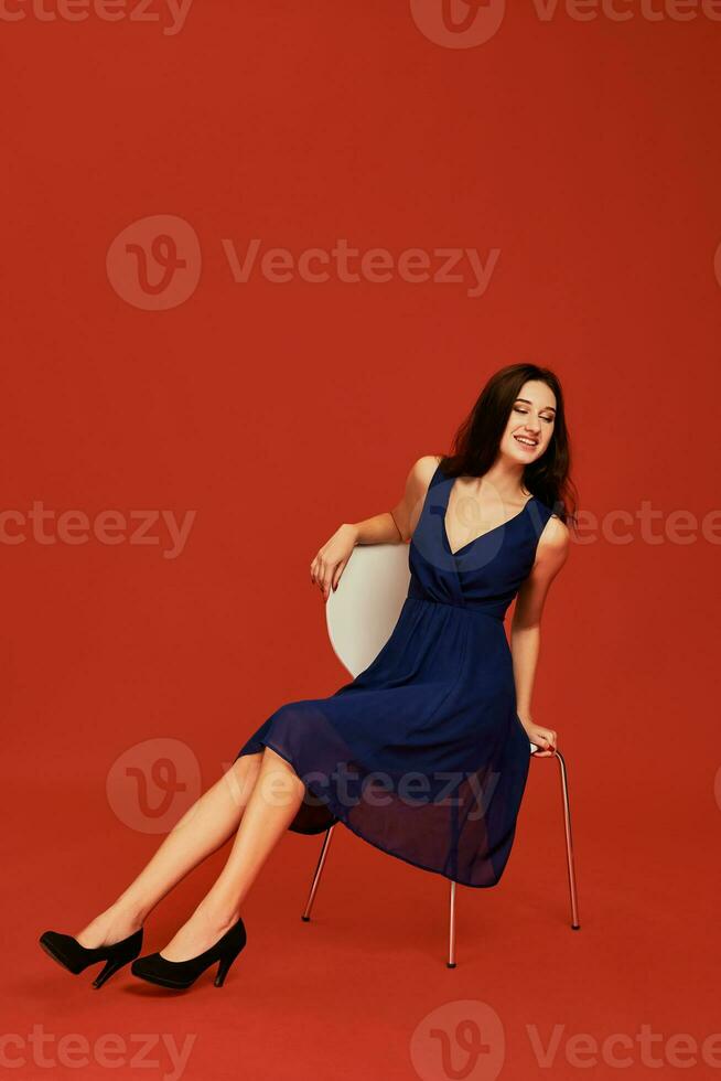 skön ung brunett kvinna i elegant blå cocktail klänning och svart hög hälar är Framställ för de kamera Sammanträde på de stol foto