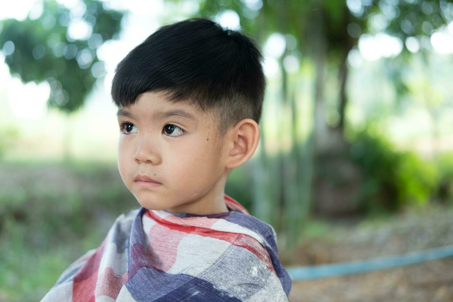 barberare skärande hår av ett asiatisk pojke i ett öppen Plats fylld med träd. foto