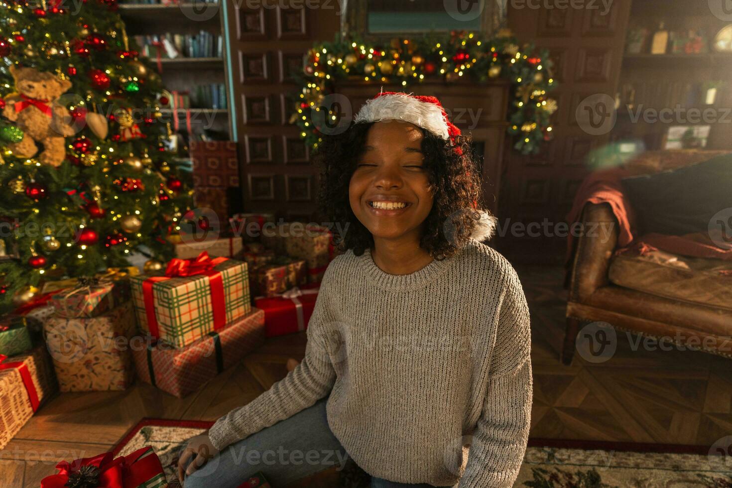 glad jul. afrikansk amerikan kvinna leende nära jul träd i klassisk mörk interiör. Lycklig flicka i santa hatt i levande rum med öppen spis jul träd gåva lådor. jul eve på Hem. foto