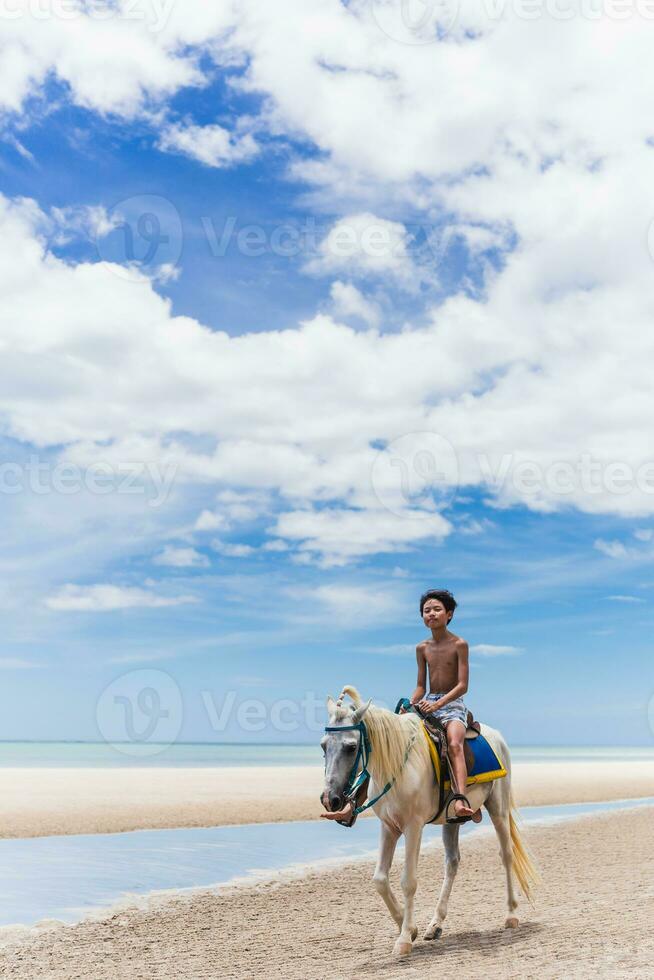 ung pojke ridning häst på de strand med blå himmel och hav i bakgrund. foto