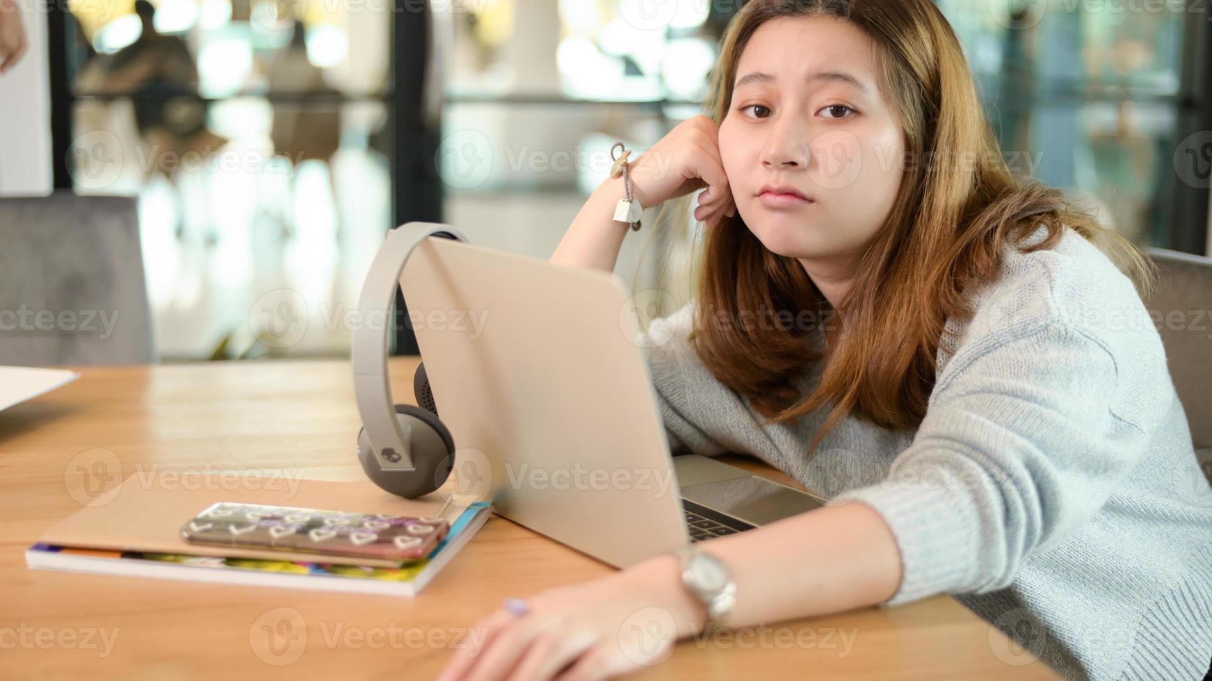 ung asiatisk tjej som tittar på kameran som får ett förvirrat ansikte att luta sig tillbaka och koppla av medan han studerar online hemma. foto