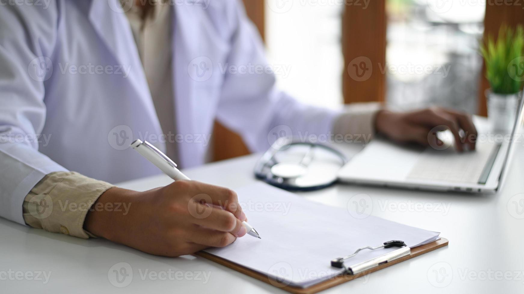medicinska koncept, läkare som bär en labrock som registrerar resultaten av auskultation på ett klippbord och bärbar dator. foto