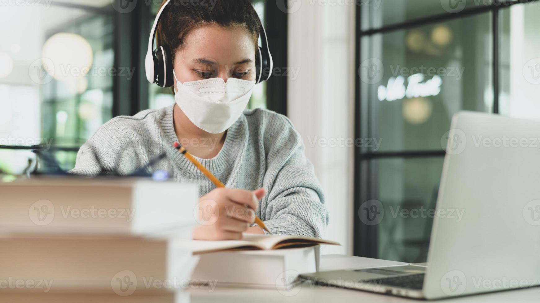 asiatisk tjej med stetoskop bär medicinsk mask som tar anteckningar och studerar online med bärbar dator. foto