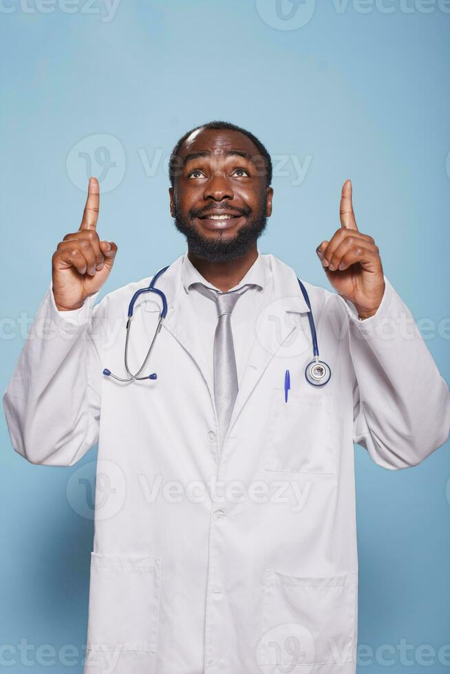 mot isolerat bakgrund, leende medicinsk läkare poäng både index fingrar upp medan bär labb täcka och stetoskop. optimistisk manlig läkare framställning en hoppfull hand gest mot de himmel. foto