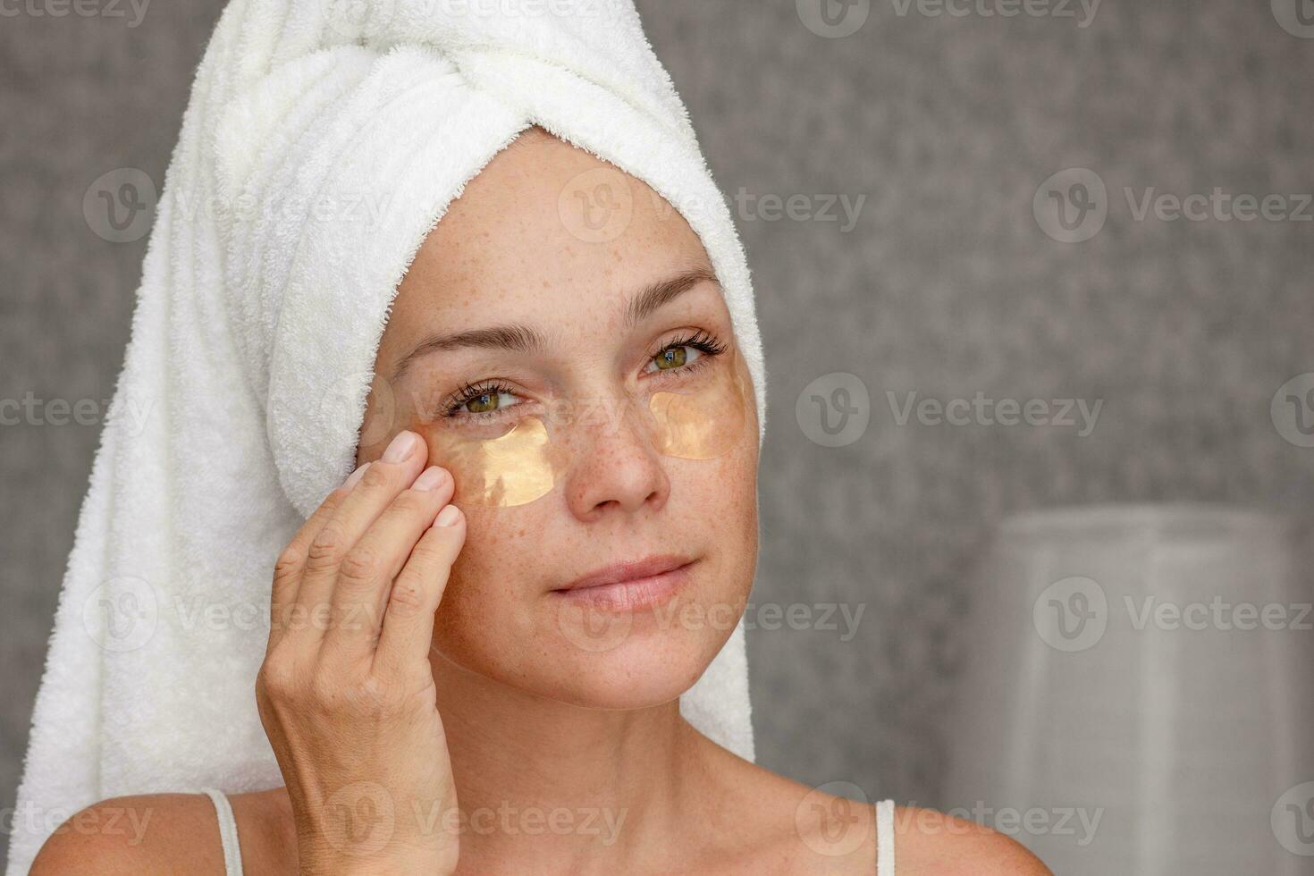 porträtt av mitten åldrig kvinna av 40s efter dusch med handduk på huvud med guld plåster under ögon på ansikte foto