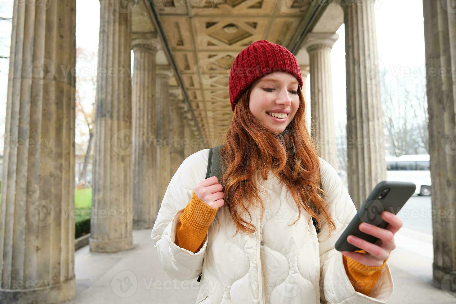 mobil bredband och människor. leende rödhårig 20s flicka med ryggsäck, användningar smartphone på gata, innehar mobil telefon och utseende på Ansökan foto