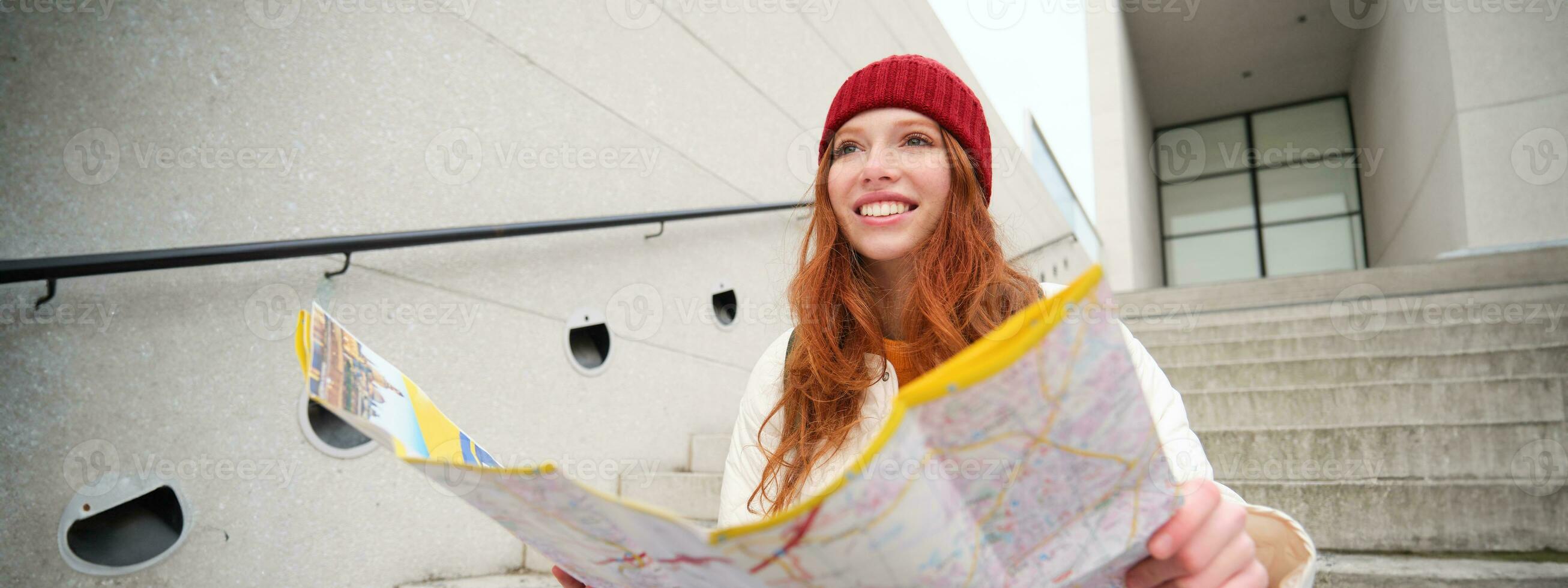 ung leende rödhårig flicka, turist sitter på trappa utomhus med stad papper Karta, ser för riktning, resande backpacker utforskar stad och utseende för sightseeing foto