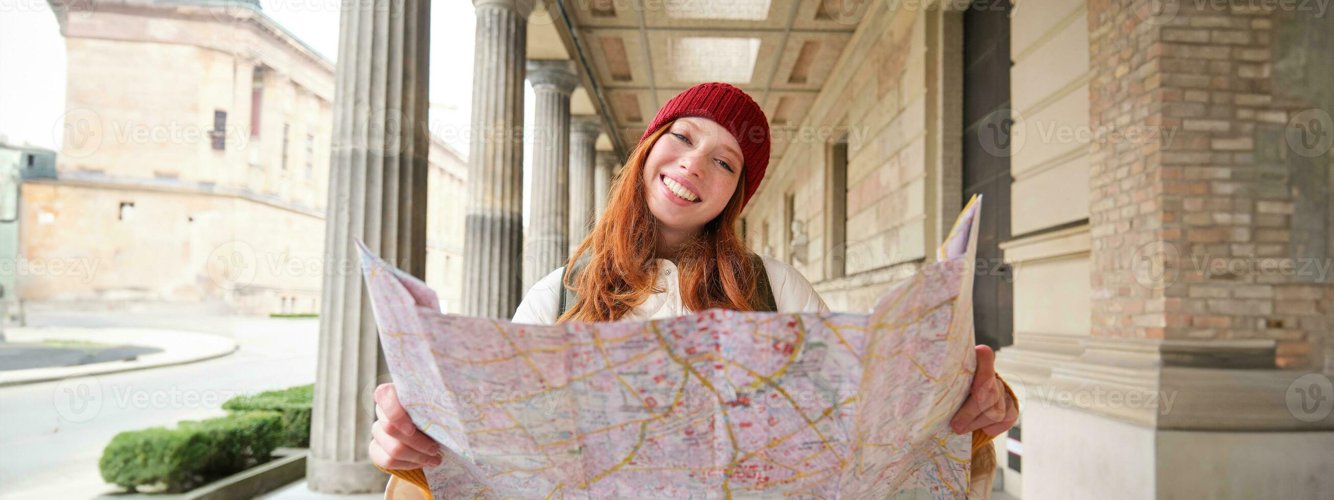 äventyrlig rödhårig flicka promenader i stad med papper Karta, utforskar stad som turist, utseende för populär turism attraktioner, utseende runt om upphetsad och ler foto