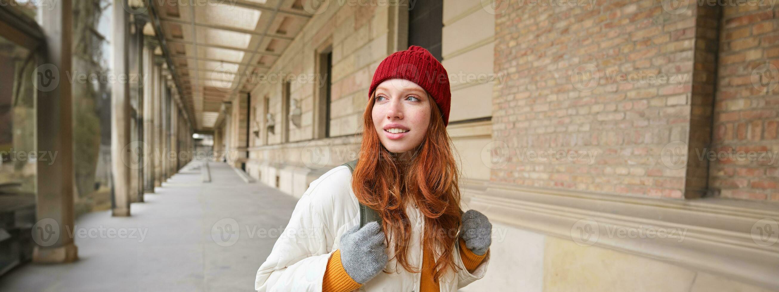 ryggsäck turist. Lycklig rödhårig flicka promenader i stad med en väska, går på resa, utforskar stad, bär röd hatt och handskar foto