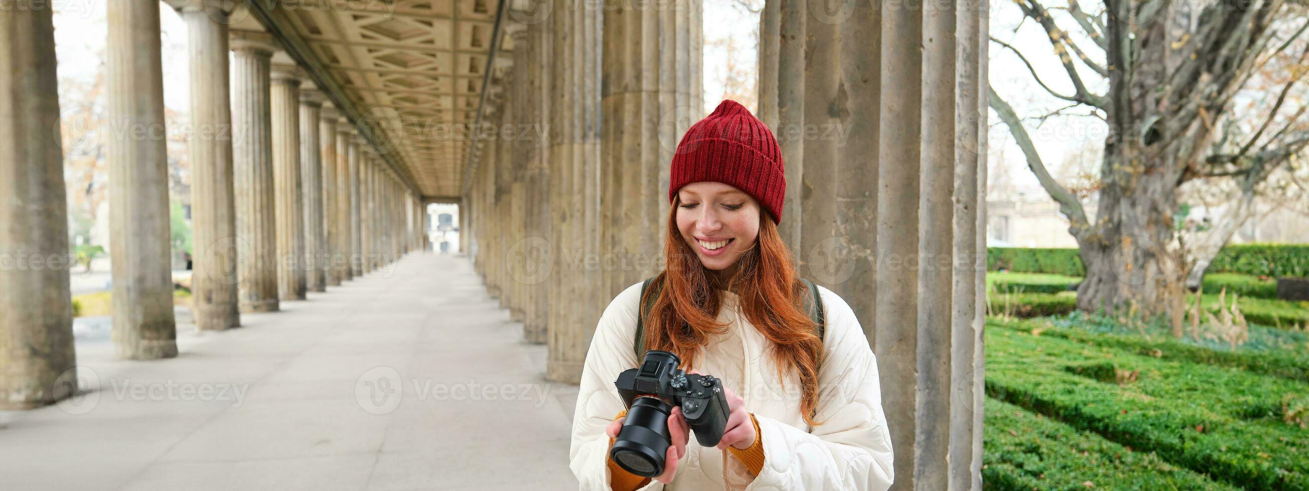 ung rödhårig kvinna fotograf, gör livsstil skytte i stad Centrum, tar foton och ler, utseende för perfekt skott, gör bild