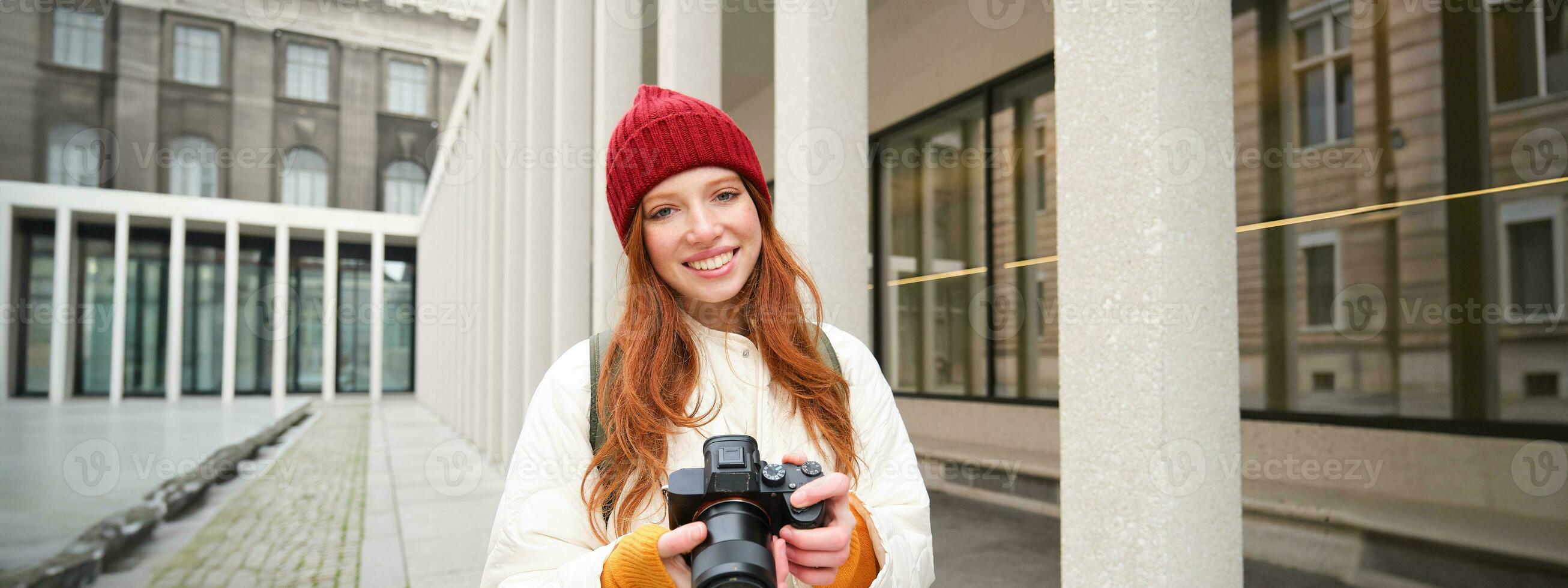 leende rödhårig flicka fotograf, tar bilder i stad, gör foton utomhus på professionell kamera