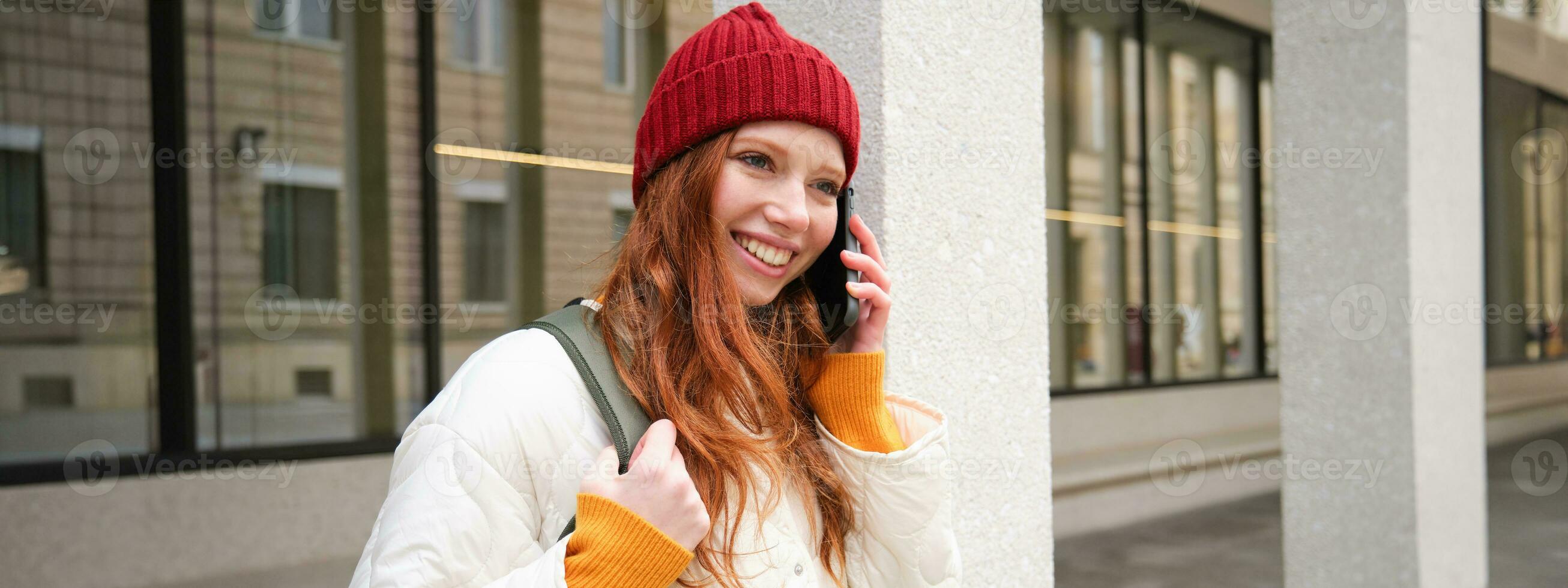 porträtt av leende ingefära flicka med röd hatt, står på gata med ryggsäck, ringar någon på telefon app, samtal på mobil, användningar smartphone foto