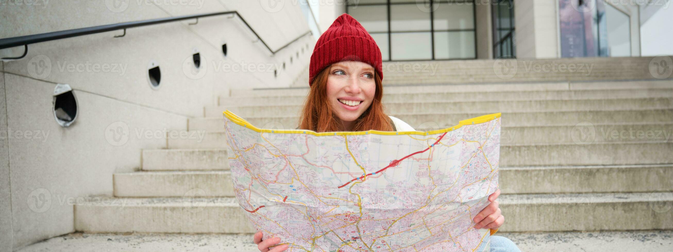 ung leende rödhårig flicka, turist sitter på trappa utomhus med stad papper Karta, ser för riktning, resande backpacker utforskar stad och utseende för sightseeing foto