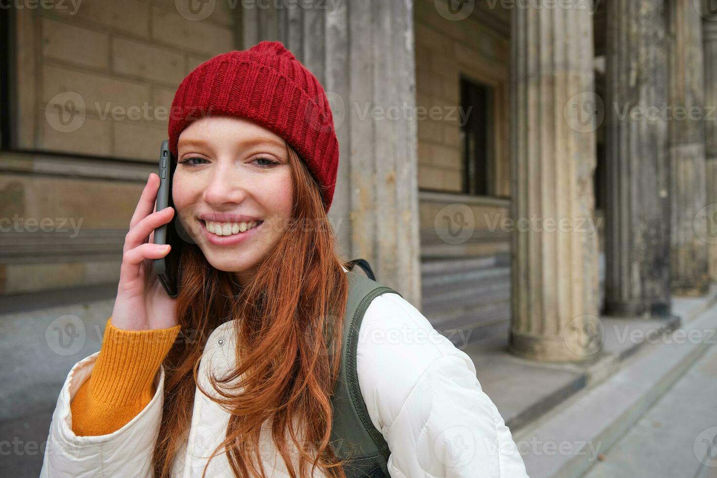 leende rödhårig kvinna turist samtal på mobil telefon och promenader runt om stad. Lycklig studerande i röd hatt samtal vän, står på gata och användningar smartphone foto