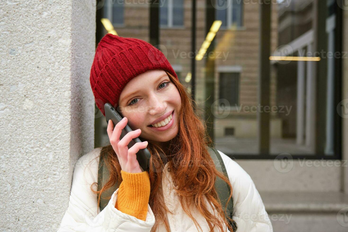 Lycklig skön flicka, rödhårig turist samtal på mobil telefon, framställning en ring upp utomlands, har konversation på smartphone, stående med ryggsäck utomhus foto