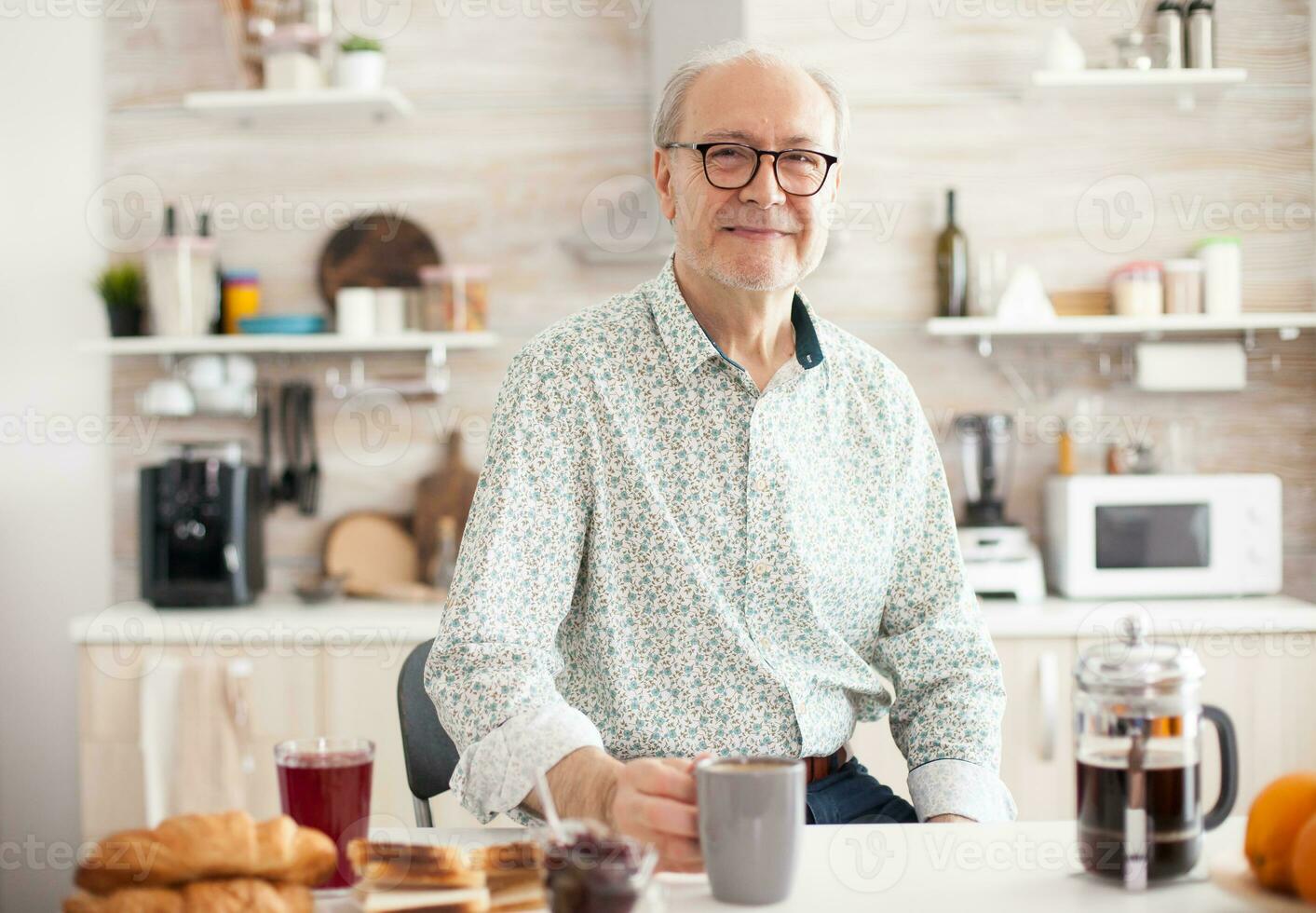 senior man i kök leende ser på kamera innehav varm kaffe kopp. porträtt av avslappnad äldre äldre person i de morgon, njuter färsk värma dryck. friska leende vuxen ansikte foto