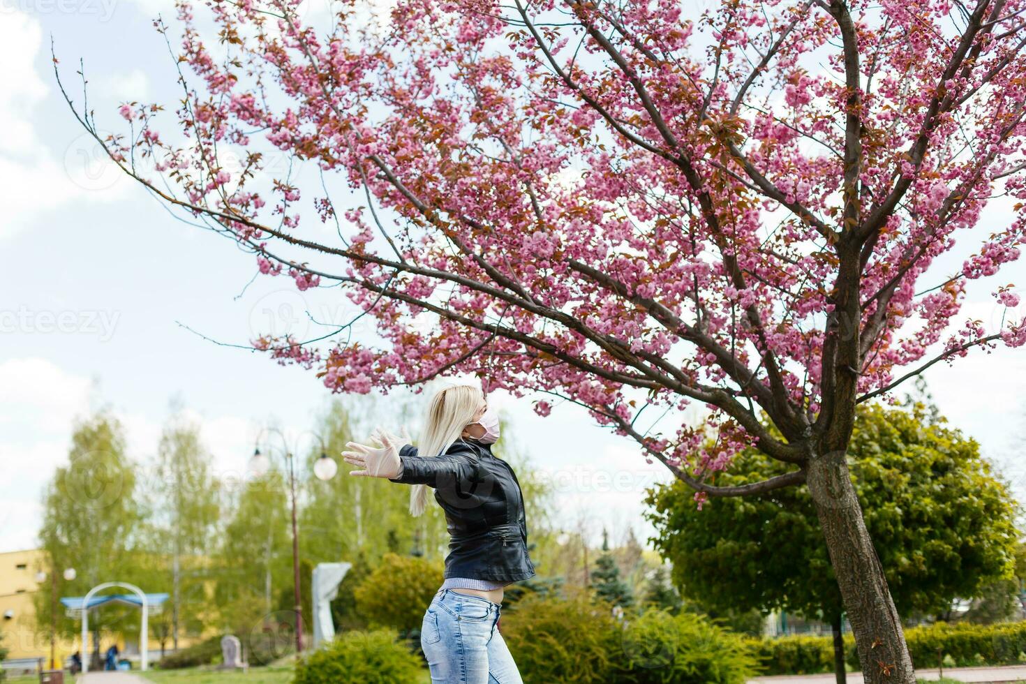 Lycklig ung kvinna bär ansiktsbehandling mask för virus skydd stående utomhus på solig vår dag. foto