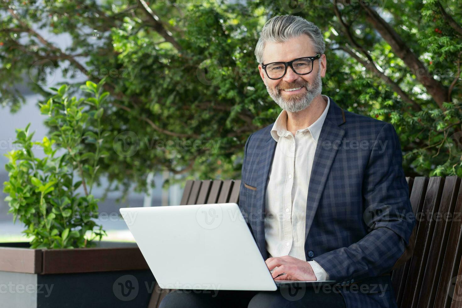 porträtt av senior affärsman utomhus i parkera på bänk, mogna chef leende och ser på kamera, man i kostym arbetssätt på bärbar dator avlägset på solig dag. foto
