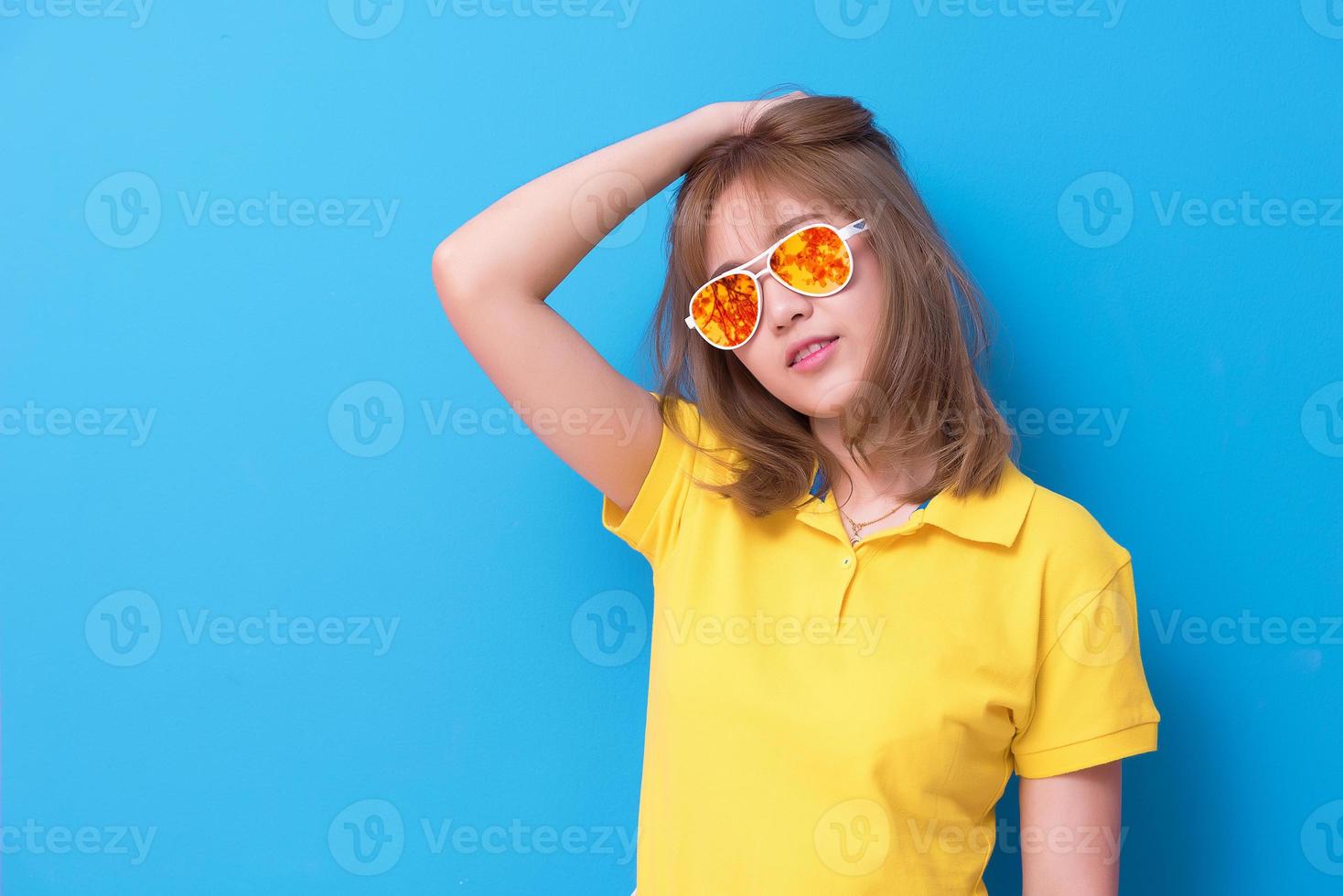 asiatisk kvinna mode poserar med mode glasögon på den blå bakgrunden. kvinna iklädd gul pikétröja och gula orange glasögon. skönhet och modernt modekoncept. foto
