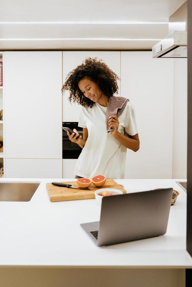 svart ung kvinna som gör frukost medan hon använder mobiltelefon i köket foto