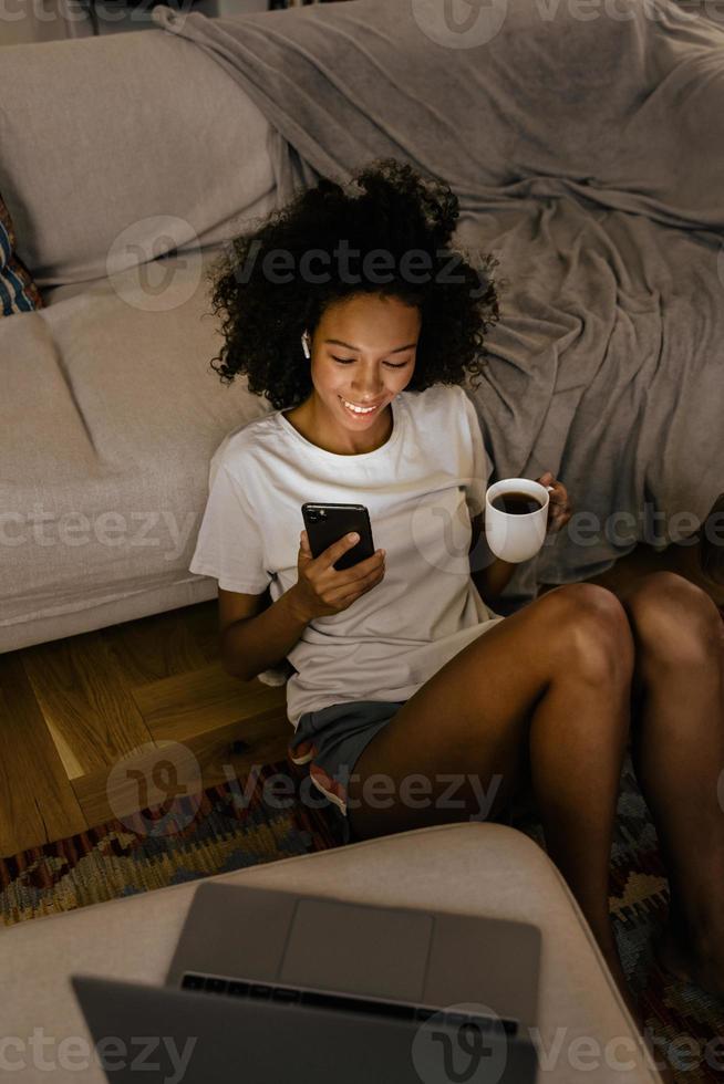 svart ung kvinna i hörlurar som använder mobiltelefon och dricker kaffe foto