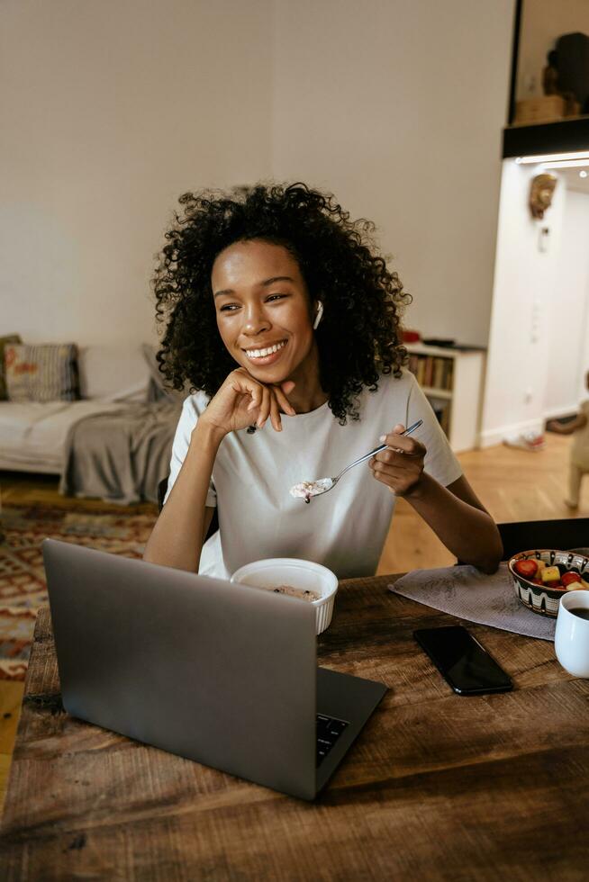 svart ung kvinna i hörlurar som använder bärbar dator och äter frukost foto