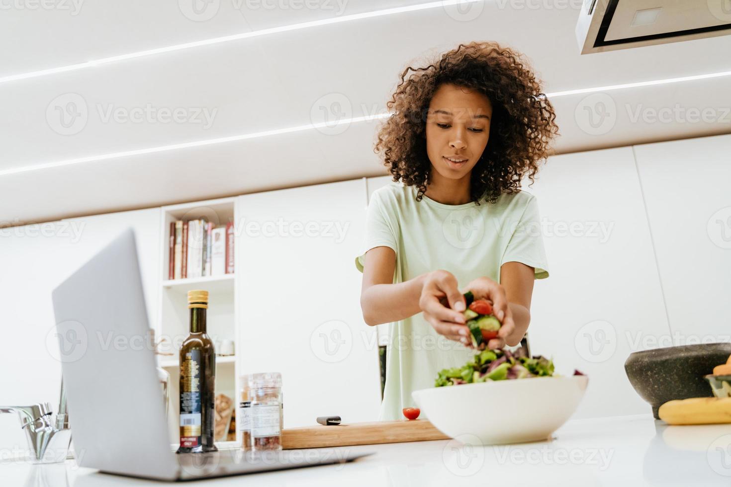 svart ung kvinna som gör sallad medan han använder bärbar dator i köket foto