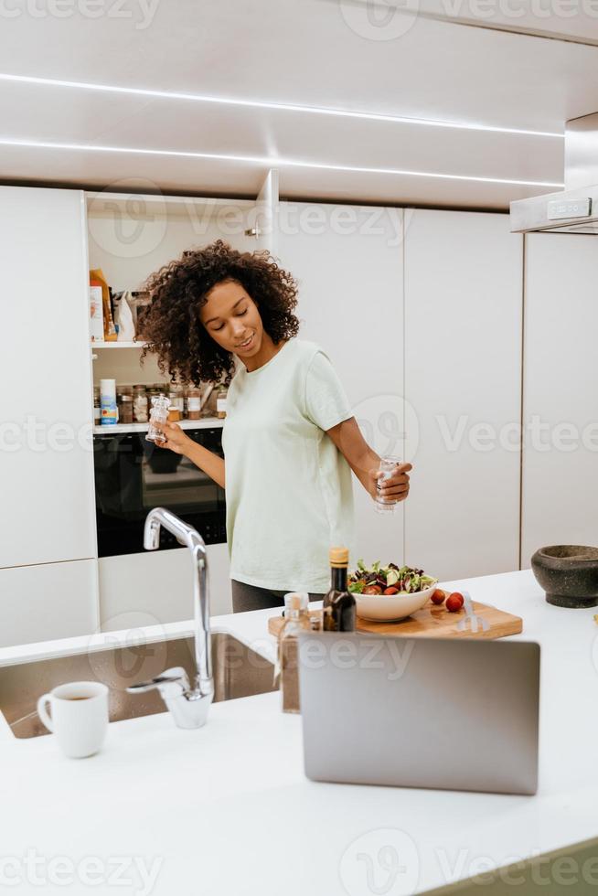 svart ung kvinna som gör sallad medan han använder bärbar dator i köket foto