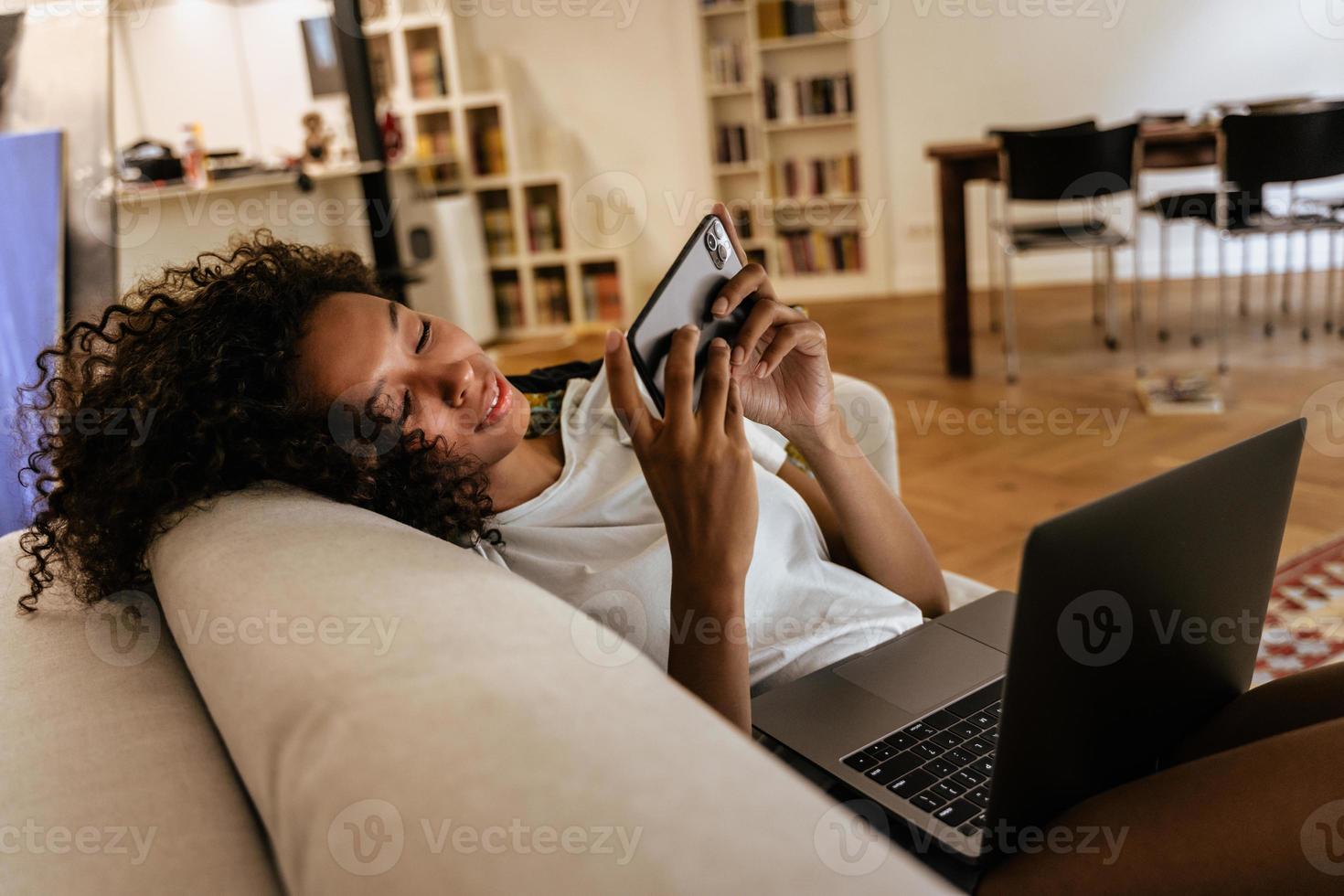 svart ung kvinna som använder mobiltelefon och laptop medan hon vilar på soffan foto