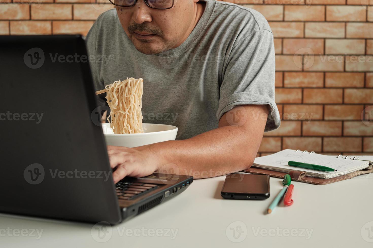 thailändsk manlig arbetare upptagen med att arbeta med bärbar dator, använd ätpinnar för att hastigt äta snabbnudlar under kontorslunchpaus, eftersom det är snabbt, gott och billigt. med tiden asiatisk snabbmat, ohälsosam livsstil. foto