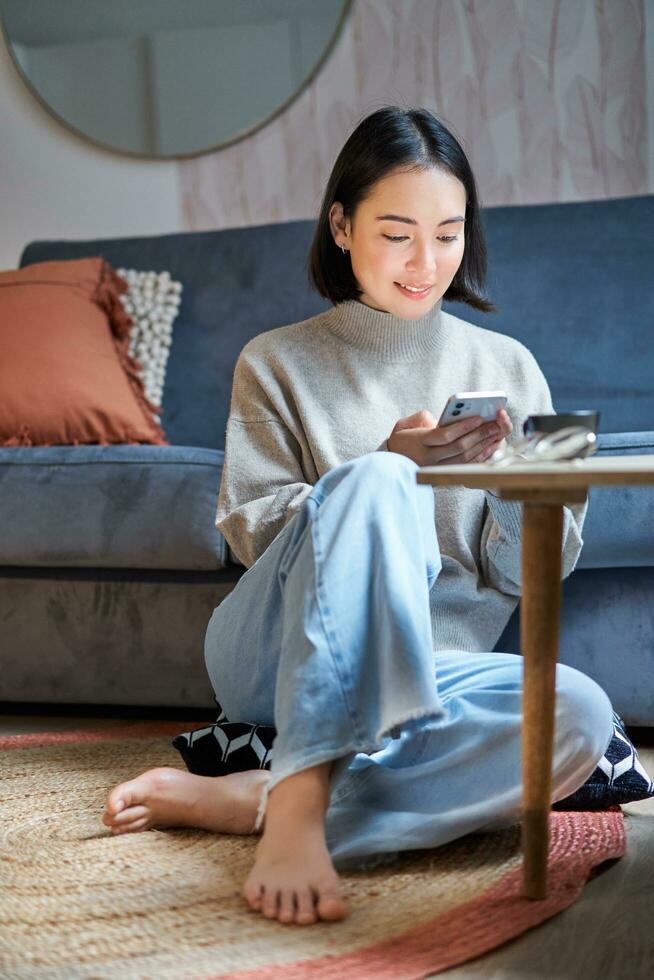 teknologi och människor. ung eleganta asiatisk kvinna sitter på Hem med henne smartphone, textning meddelande, använder sig av Ansökan på mobil telefon foto