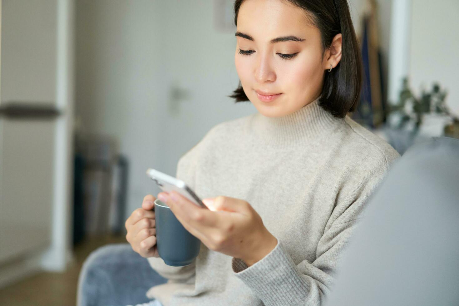 porträtt av leende asiatisk flicka kontroll henne Nyheter utfodra på smartphone och dricka kaffe, Sammanträde på soffa på Hem, bläddring på mobil telefon, läsning foto