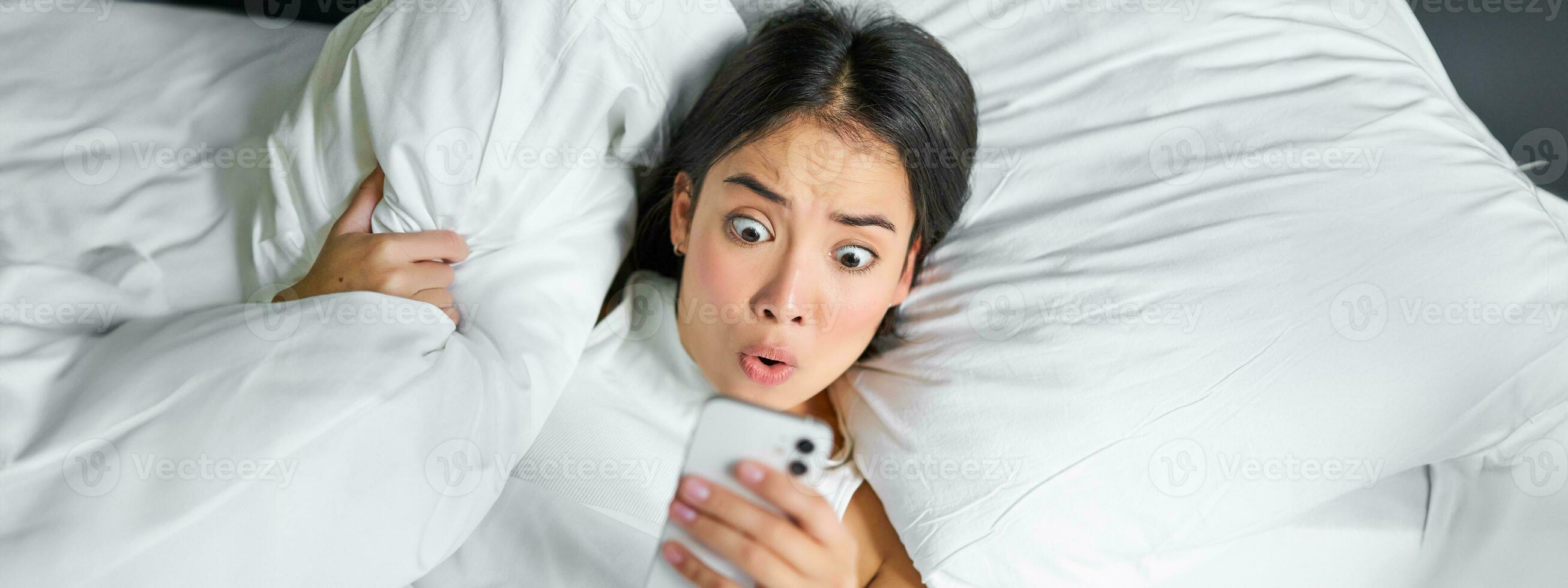 porträtt av asiatisk flicka vaknar upp i morgon, utseende på mobil telefon med chockade ansikte, försov sig, kramas kudde och grimaserande foto