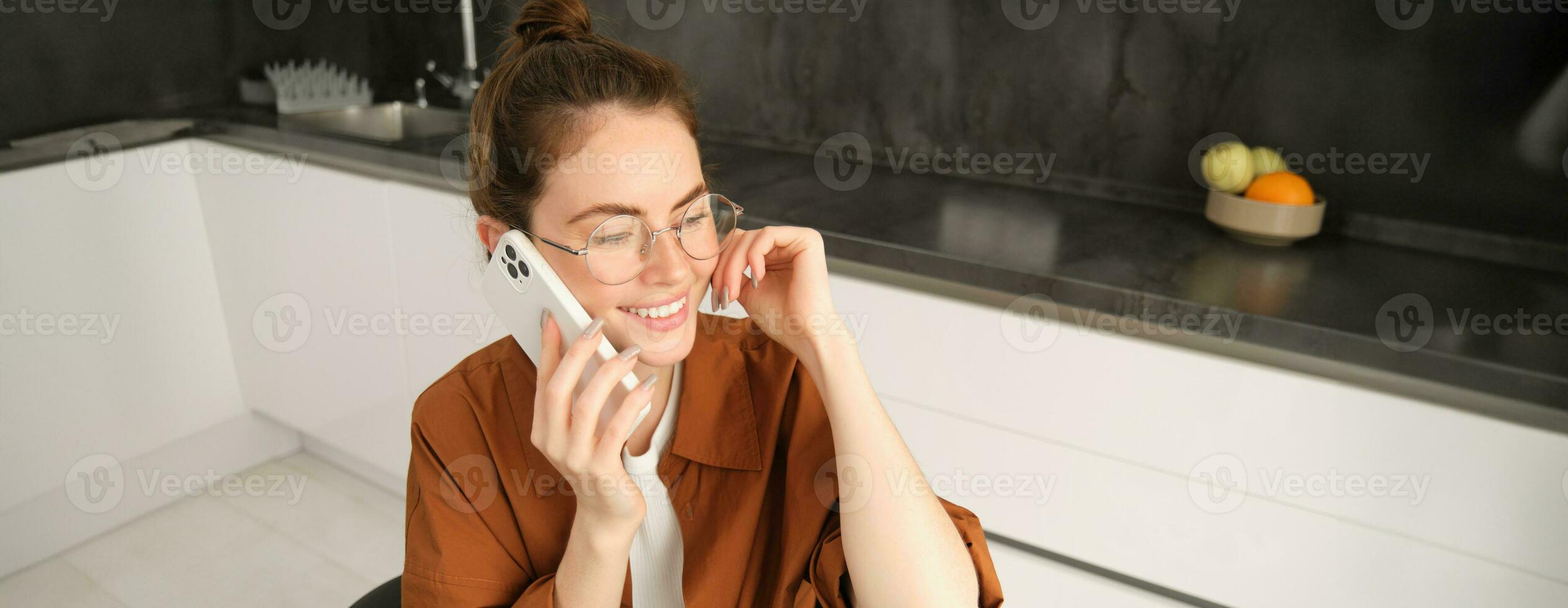 modern ung kvinna Sammanträde i kök med smartphone, talande på mobil telefon, har vänlig konversation, kallelse någon foto
