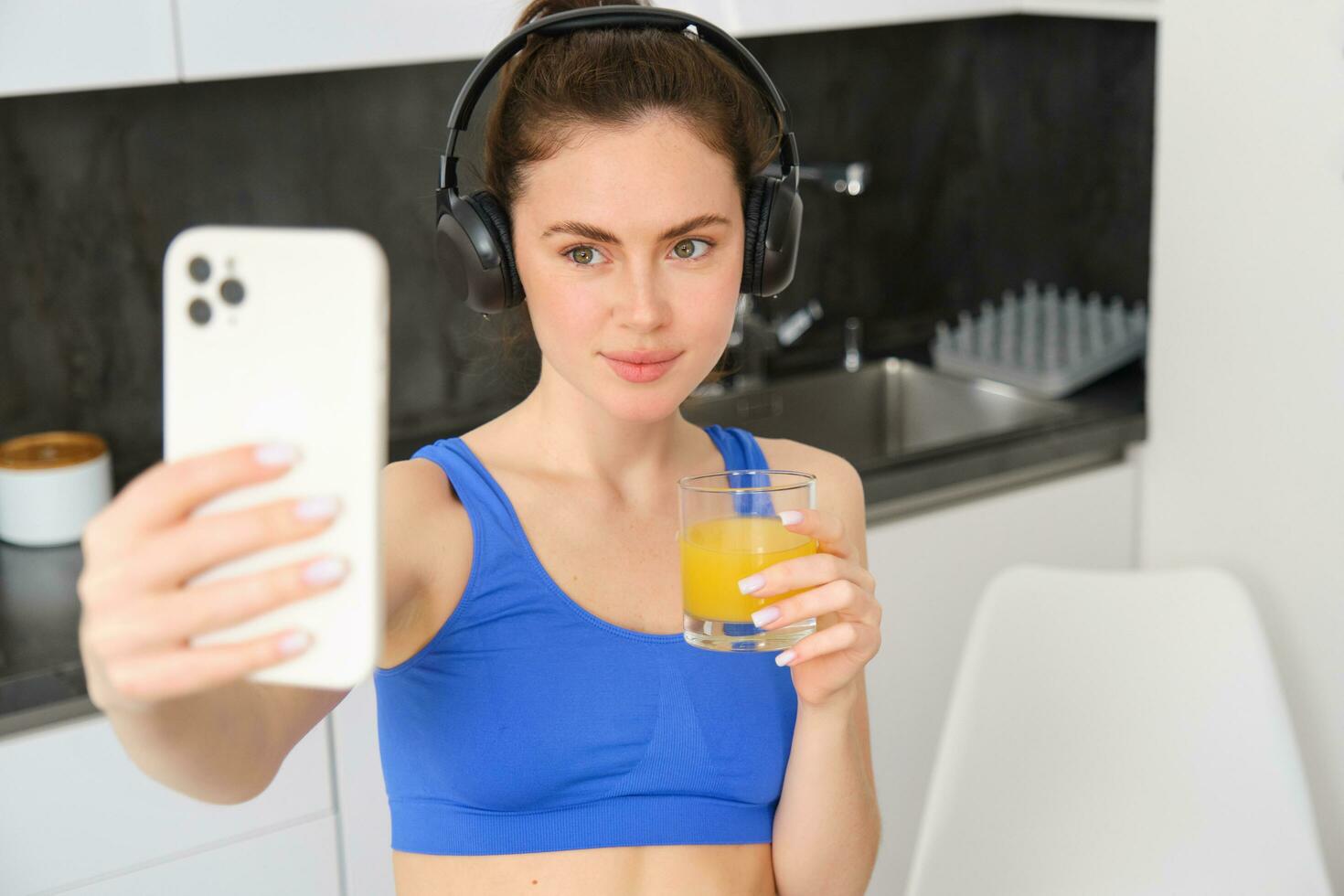 porträtt av ung livsstil, kondition bloggare, kvinna med orange juice, tar selfie på smartphone kamera, Framställ i aktiva kläder och trådlös hörlurar foto