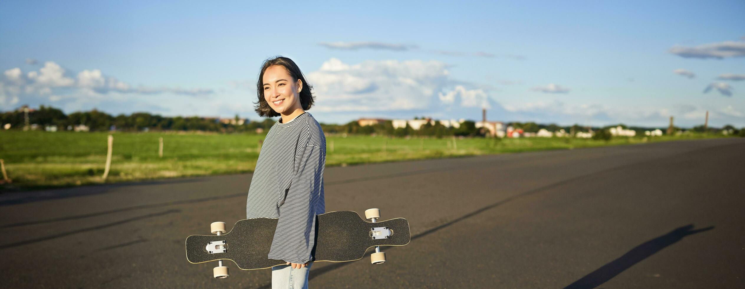 vertikal skott av sorglös asiatisk flicka med longboard. ung kvinna skater innehav kryssare på henne axlar och gående på väg, skateboard foto