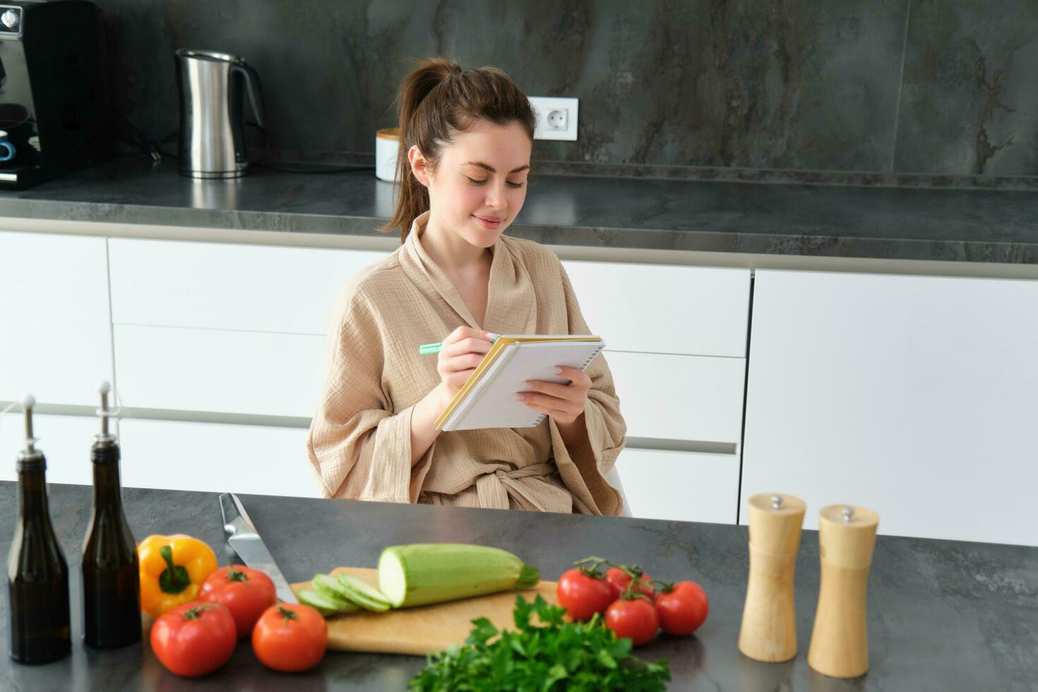 porträtt av Lycklig ung kvinna skrivningar ner meny för middag, sitter i de kök nära grönsaker, gör matvaror lista för handla, poser i de morgonrock foto