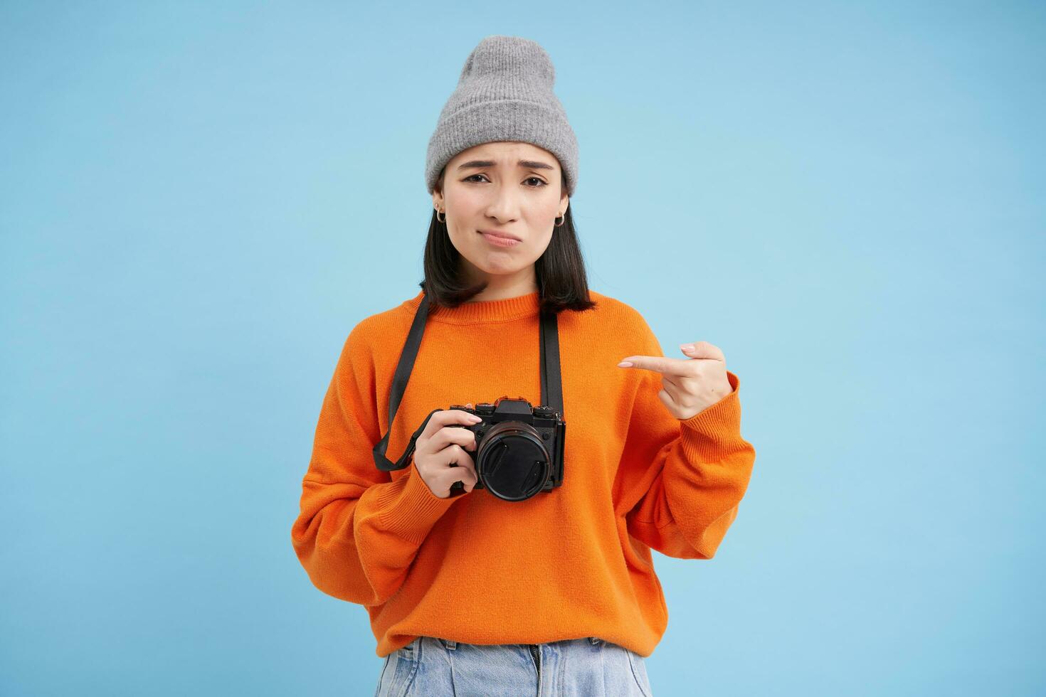 asiatisk flicka i hatt, poäng på henne digital kamera med besviken, upprörd ansikte, inte tycka om henne enhet, fotograf klagar på henne digitalkamera, blå bakgrund foto