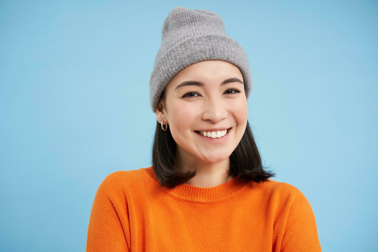 stänga upp porträtt av leende, skön ung kvinna, asiatisk flicka i hatt, ser Lycklig och uppriktig på kamera, stående över blå bakgrund foto
