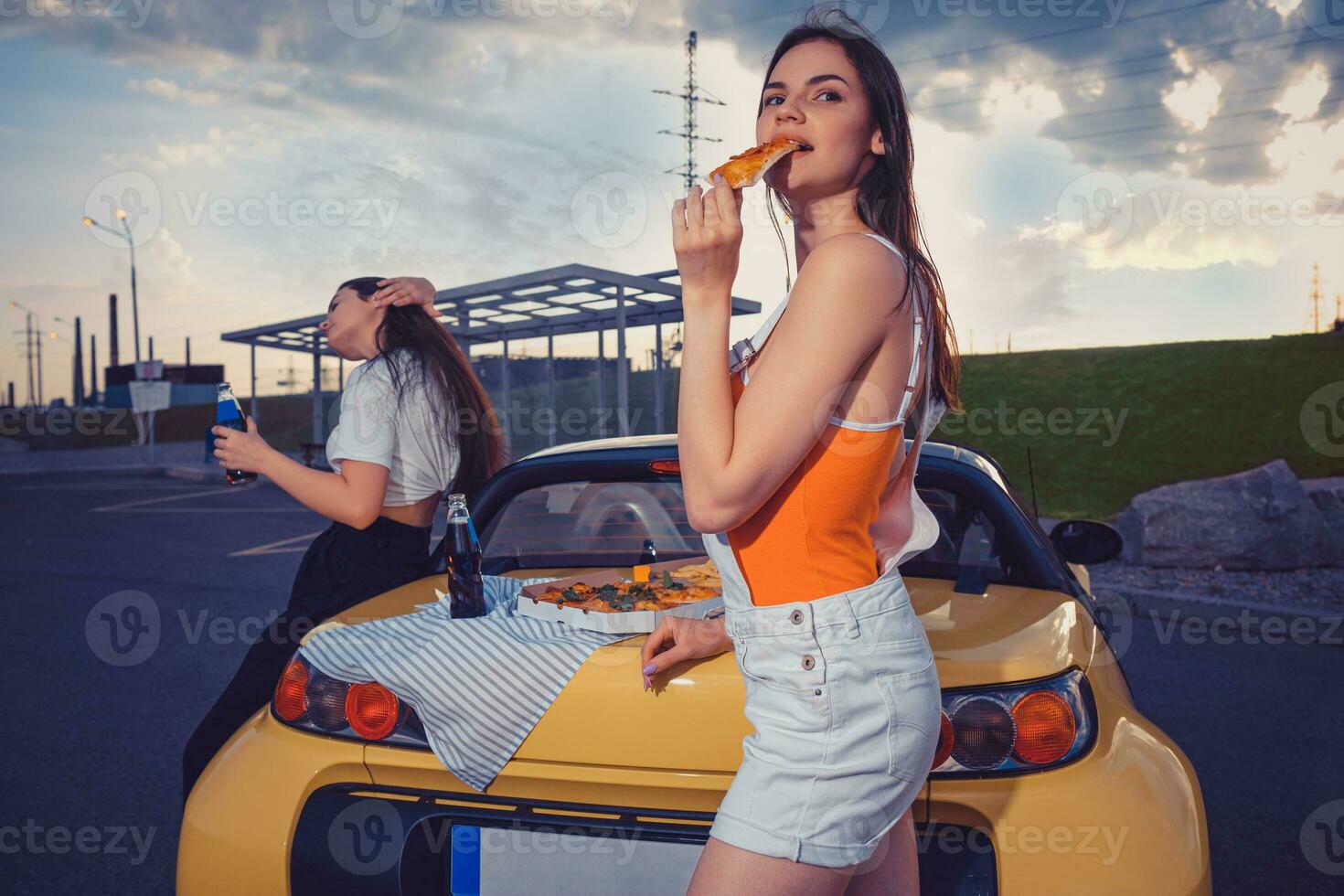 attraktiv kvinnor i tillfällig kläder äter pizza, njuter kolsyrad dryck i glas flaskor medan Framställ nära gul bil på parkering massa. kopia Plats foto